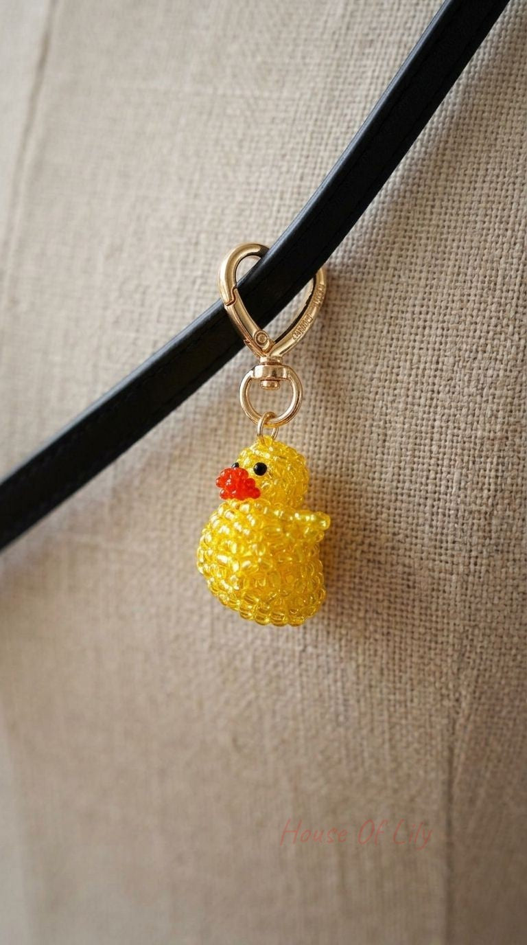 Delicate beaded duck bag charm with yellow, orange, and black beads, clipped on a black leather strap with a neutral fabric background.