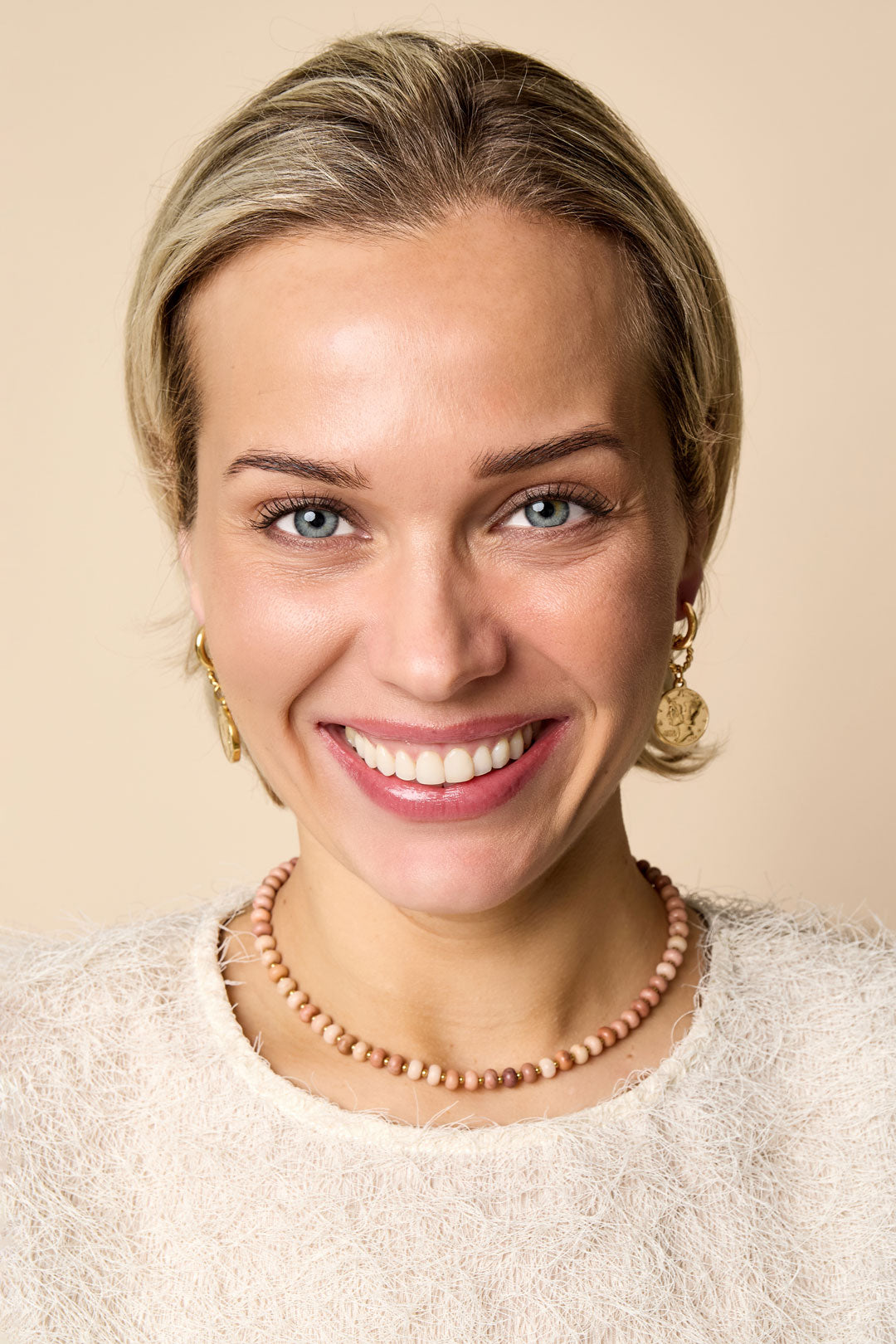 Woman smiling wearing a pink stone beaded necklace and gold coin drop earrings against beige background
