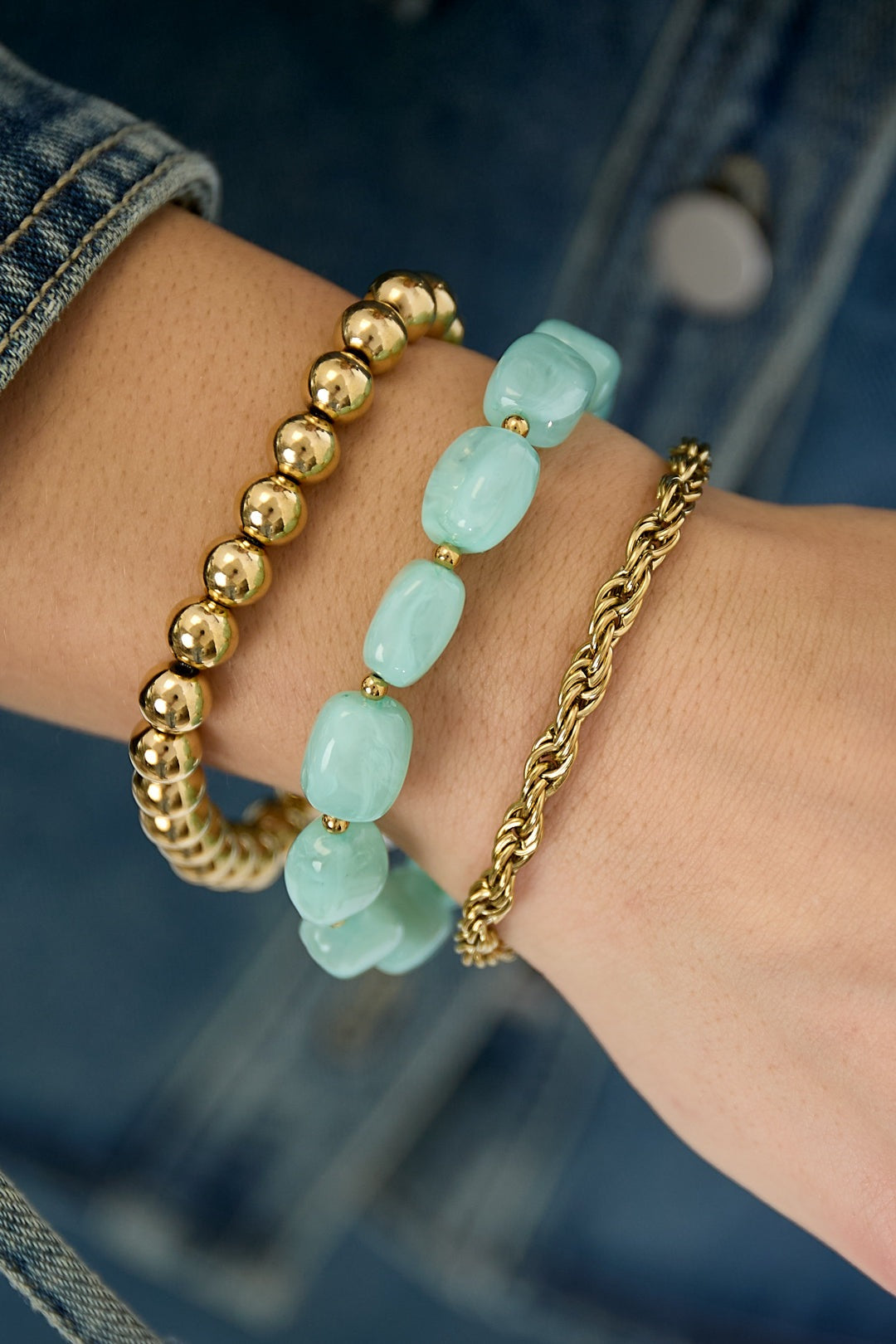 Wrist adorned with three bracelets: shiny gold-tone beads, mint green marbled acrylic beads, and a twisted gold-tone chain, with denim sleeve in background