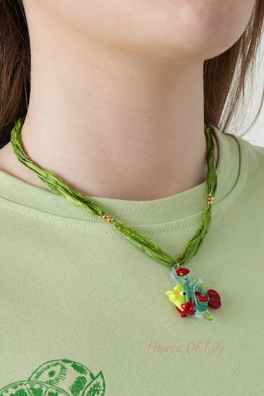 Model wearing green fabric cord necklace with colorful fish glass pendant around the neck over light green shirt