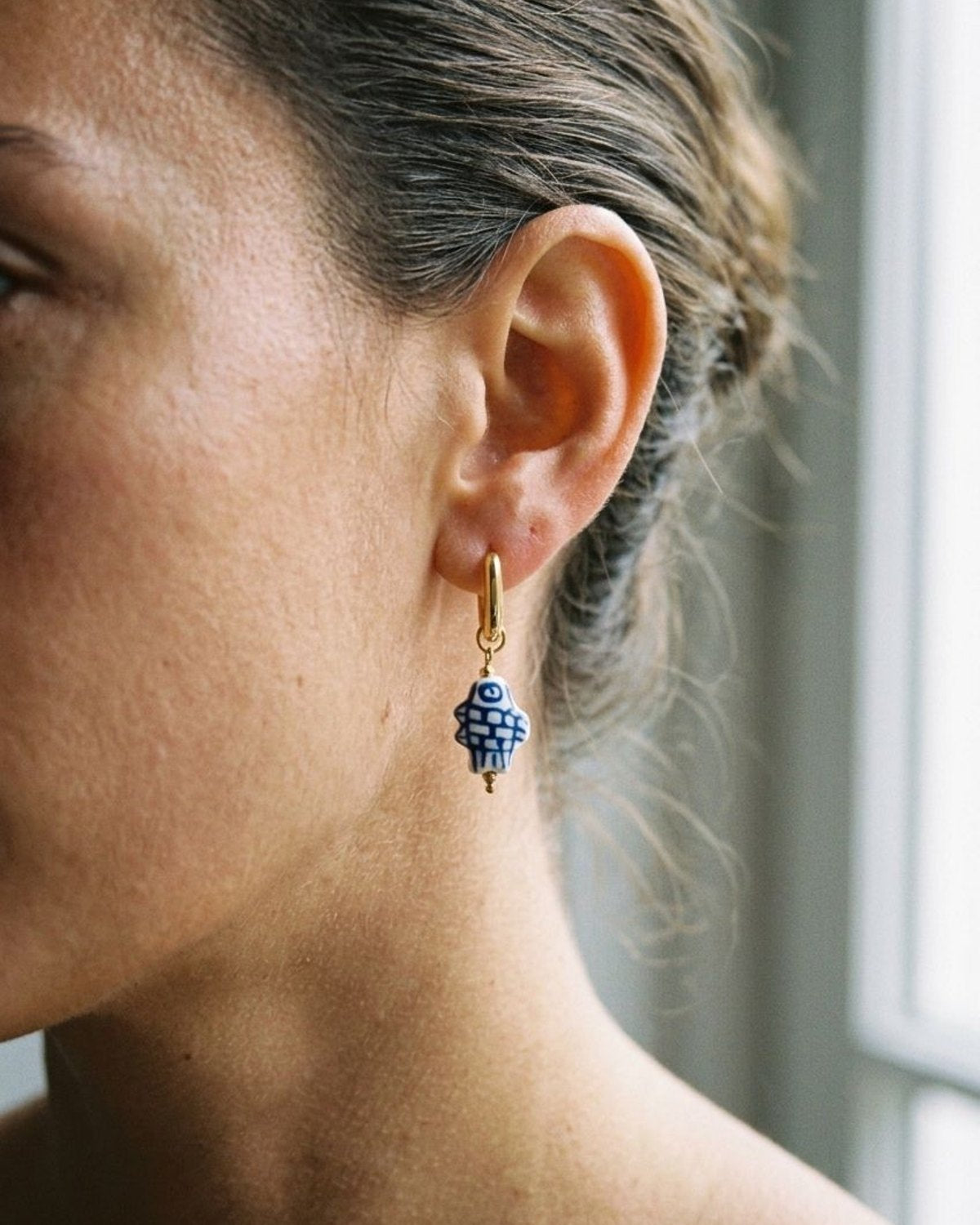 Woman wearing a delicate blue and white checkered ceramic fish earring hanging from a gold loop.