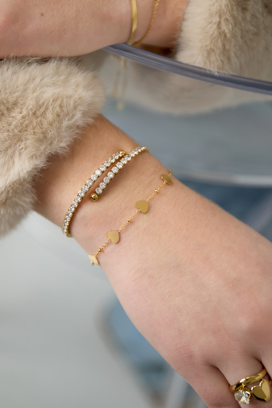 Close-up of a woman's wrist wearing the gold heart chain bracelet along with a double-row rhinestone bracelet and gold rings, styled with a cozy beige fur sleeve