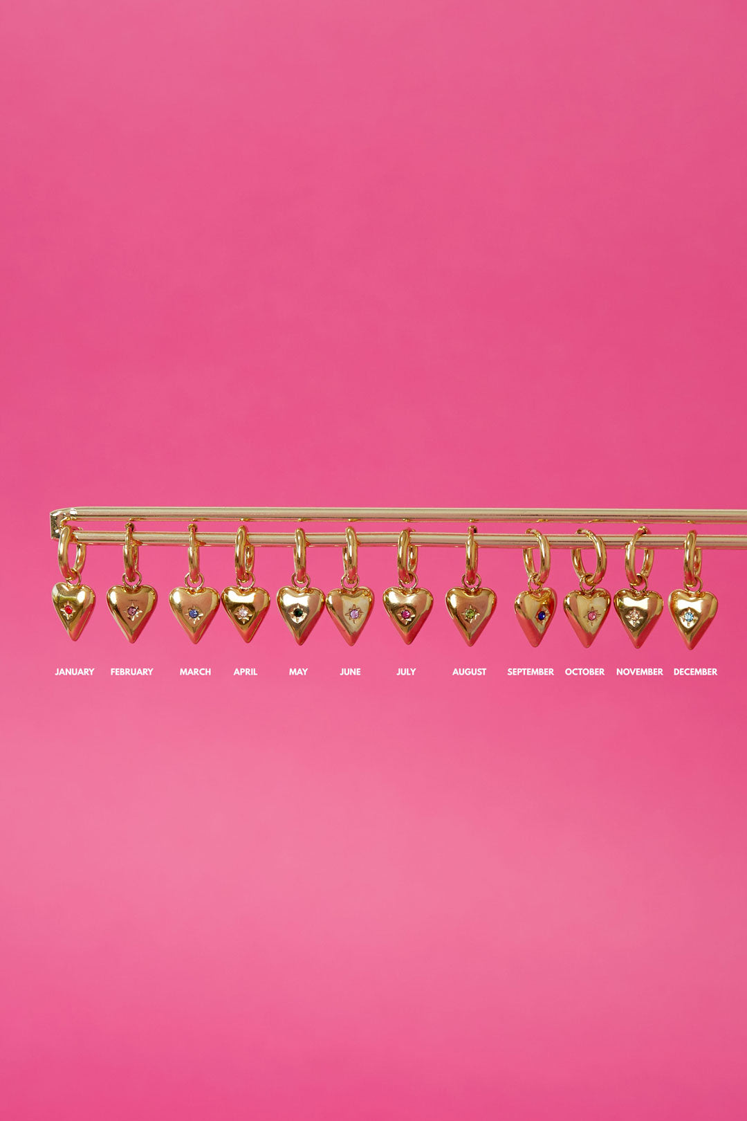 Display of twelve gold-plated heart charm birthstone pendants hanging on a horizontal bar against a pink background
