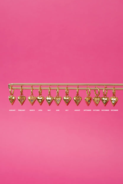Display of twelve gold-plated heart charm birthstone pendants hanging on a horizontal bar against a pink background