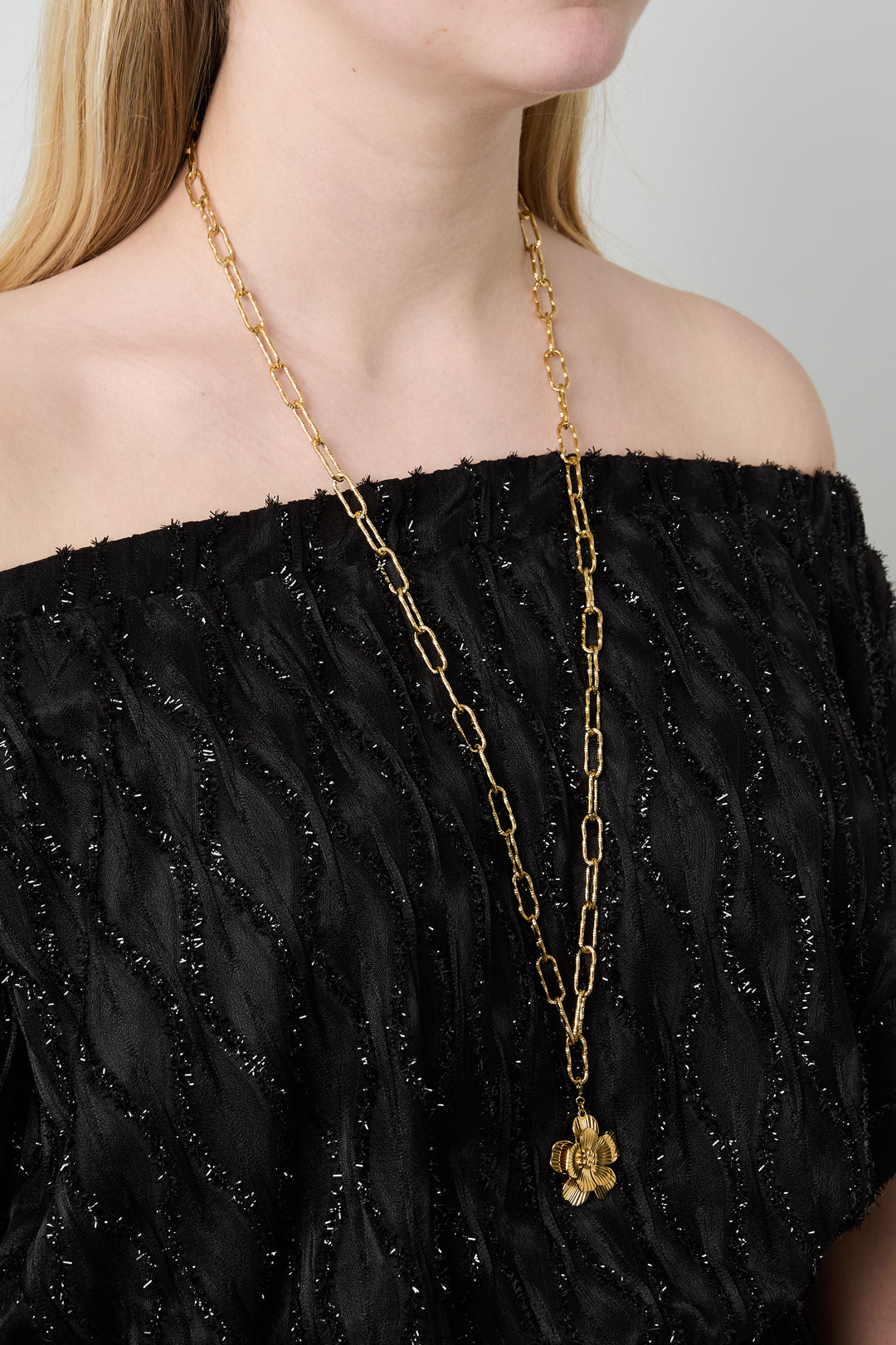 Model wearing a long gold link chain necklace with a detailed flower pendant, paired with black textured off-shoulder top