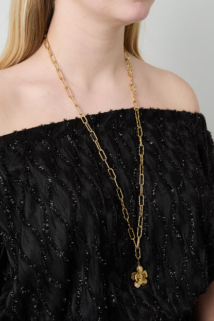 Model wearing a long gold link chain necklace with a detailed flower pendant, paired with black textured off-shoulder top
