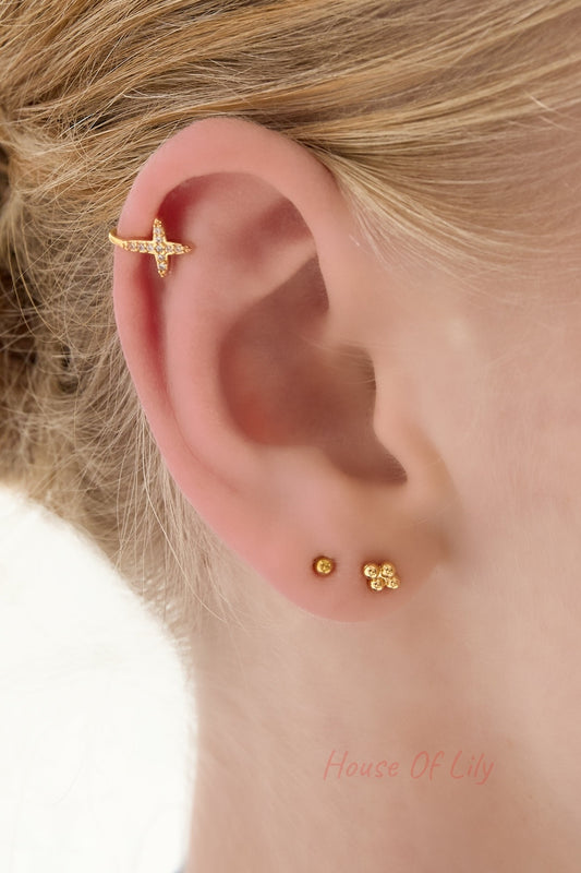 Gold ear cuff with cross-shaped cubic zirconia detail worn on the upper ear alongside small gold stud earrings