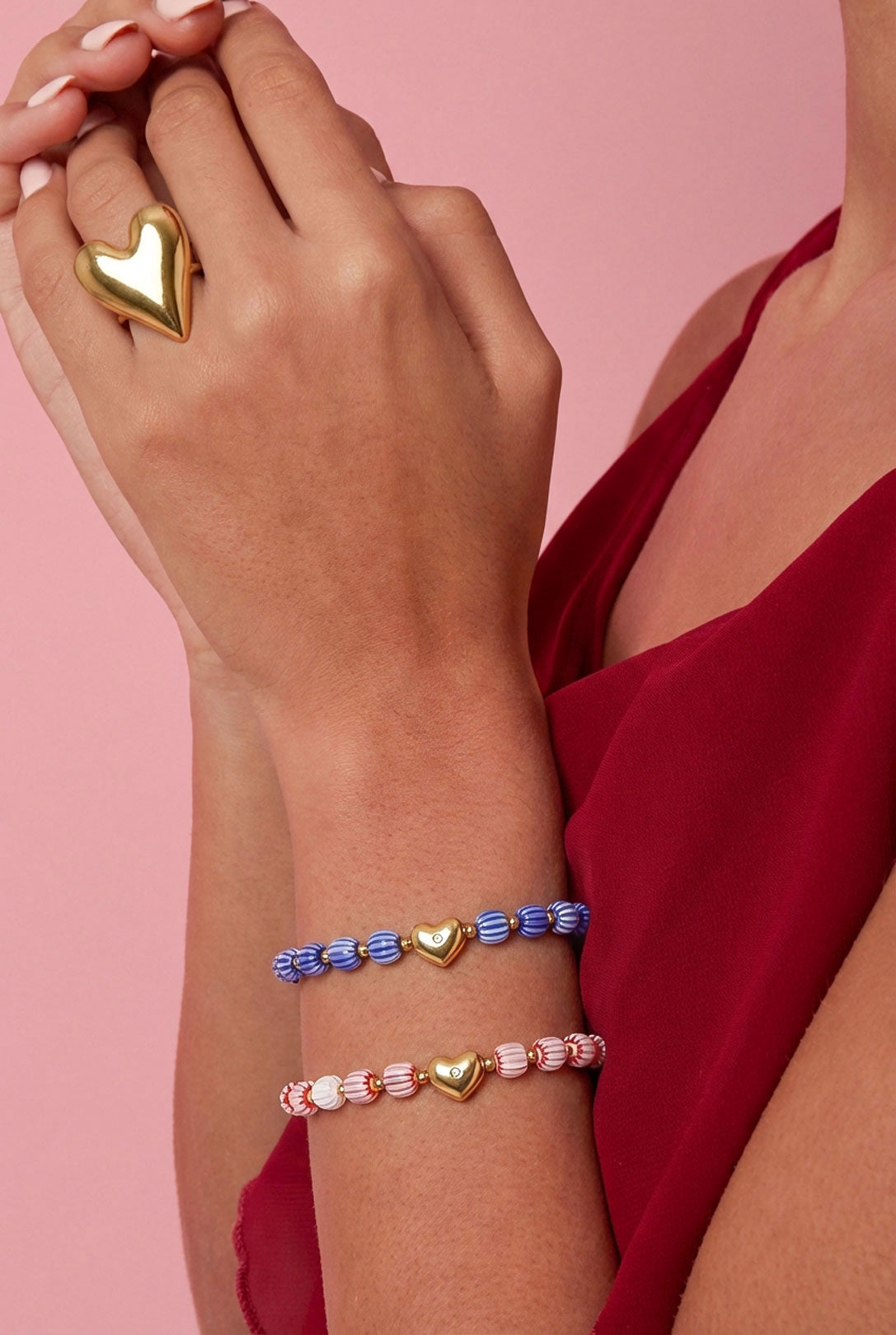 Close-up of a woman's wrist wearing blue and white, and pink and white striped beaded bracelets with gold heart charms, paired with a gold heart ring, against a pink background