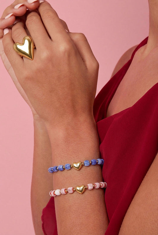 Close-up of a woman's wrist wearing blue and white, and pink and white striped beaded bracelets with gold heart charms, paired with a gold heart ring, against a pink background