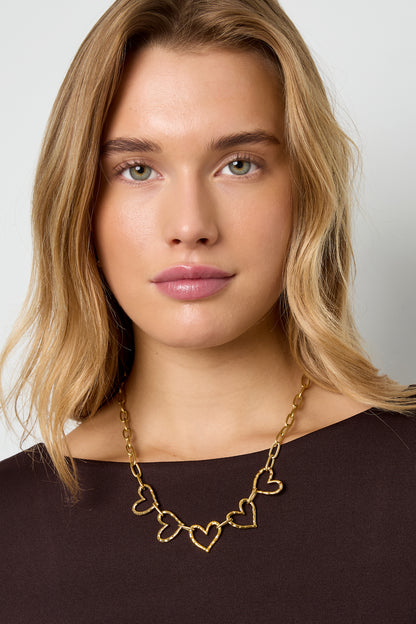 Close-up of woman wearing gold-tone heart link necklace with five hammered heart charms against dark top