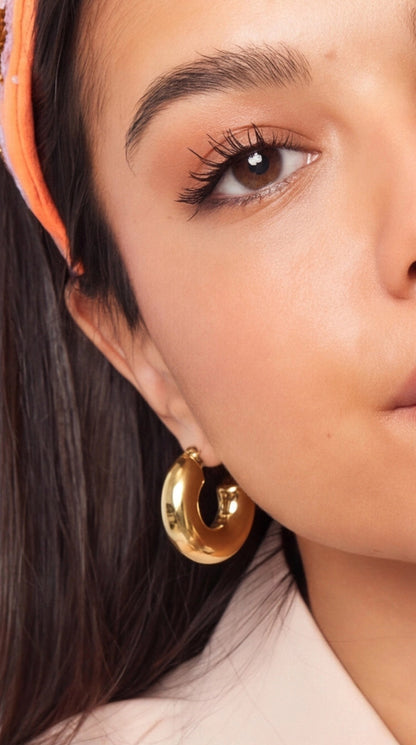 Close-up of a woman wearing thick, polished gold hoop earrings with a chunky, rounded design.