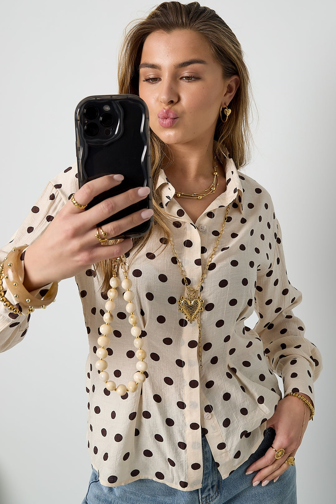 Woman taking selfie showing gold bead layered necklace and long gold chain with heart pendant over polka dot blouse