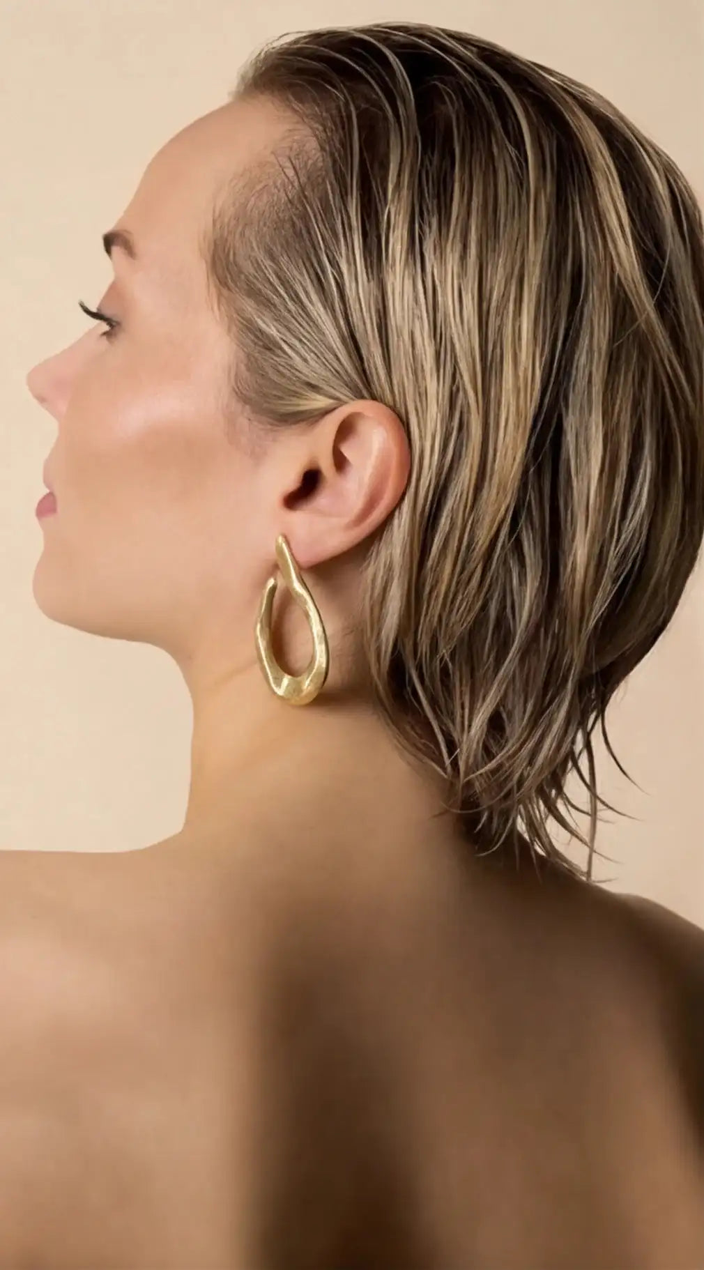 Woman with a styled haircut and gold hoop earrings against a beige background