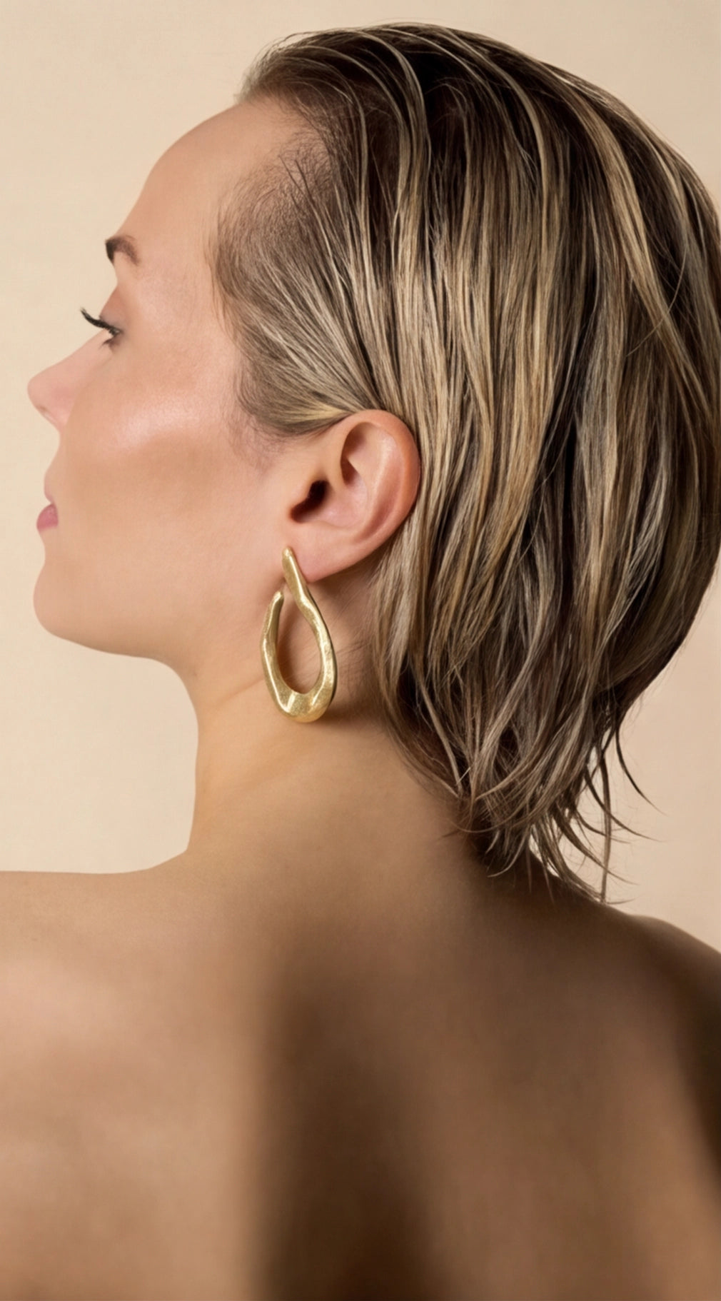 Woman with a styled haircut and gold hoop earrings against a beige background