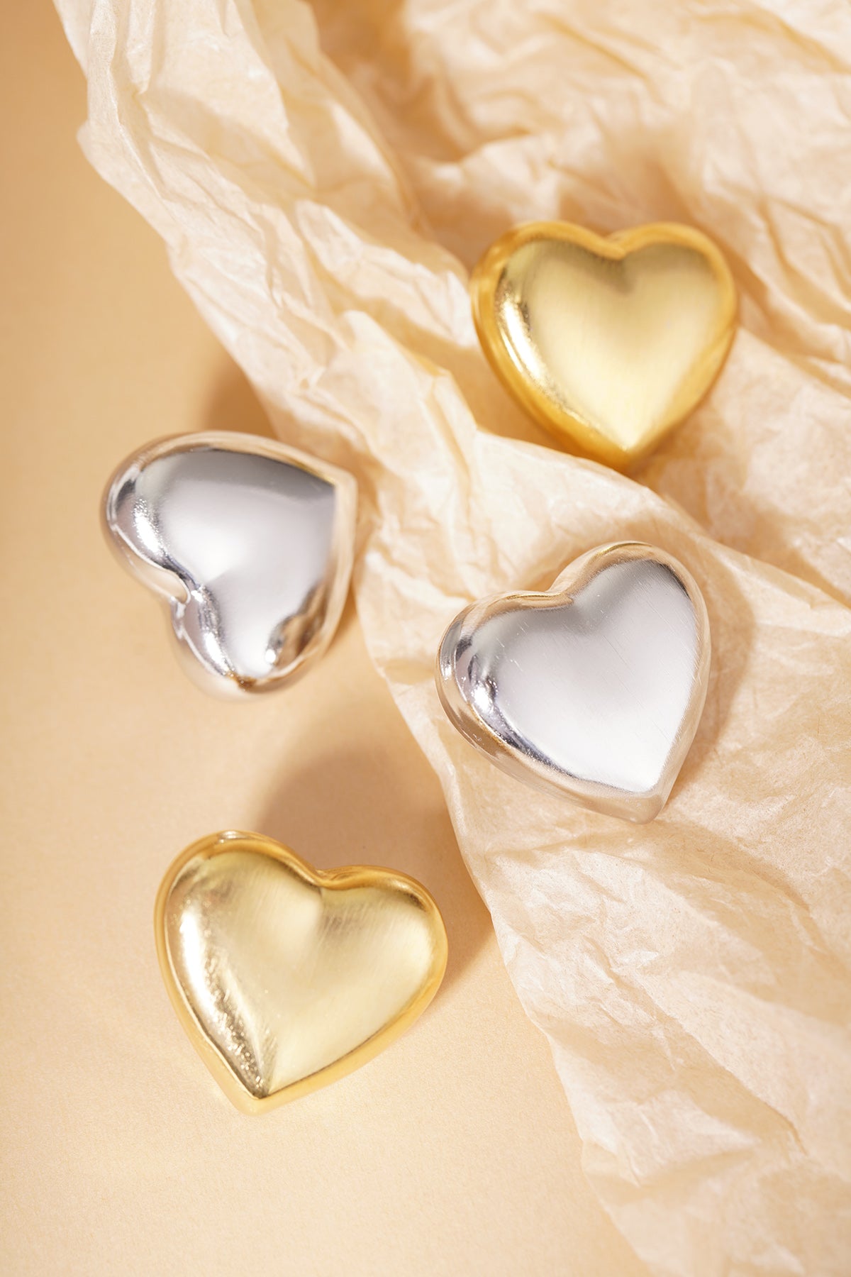 Group of matte gold and silver heart stud earrings arranged on neutral beige paper with texture