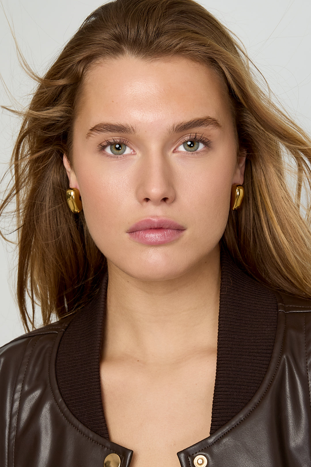 Front view of model wearing chunky gold heart hoop earrings with loose hair and brown jacket