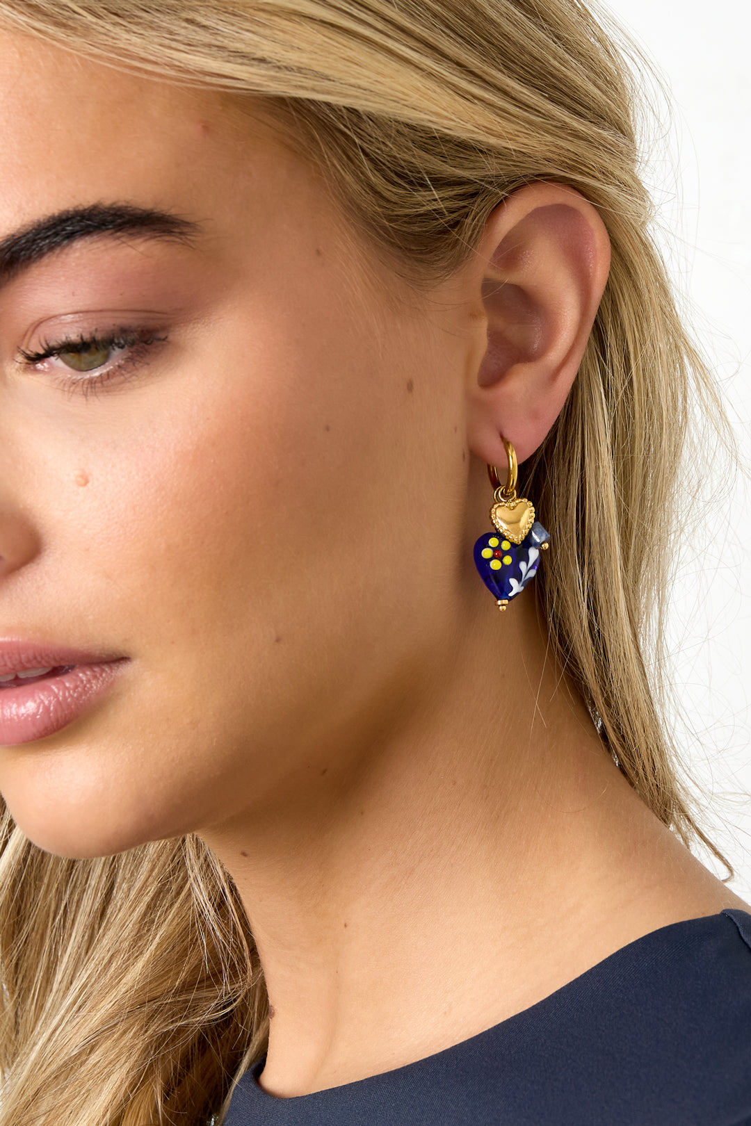 Close-up of woman wearing gold hoop earring with dark blue heart charm and gold heart pendant