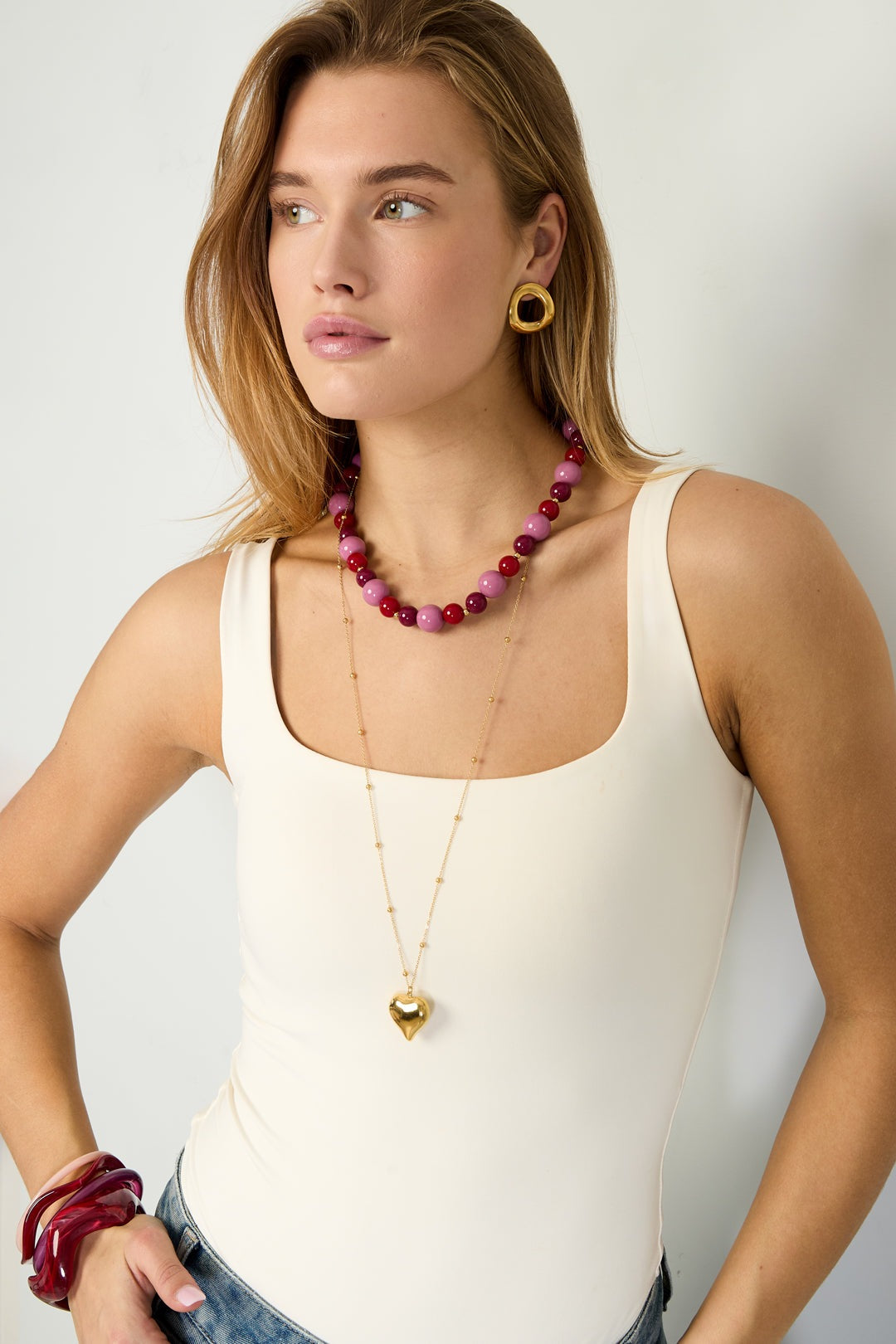Model wearing pink and red chunky beaded necklace paired with white tank top and gold heart pendant necklace
