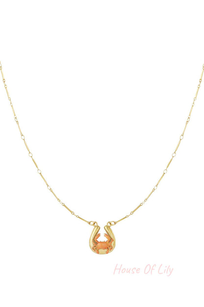Gold necklace featuring a horseshoe charm with a small, coral-colored crab, on a delicate gold bar-link chain.