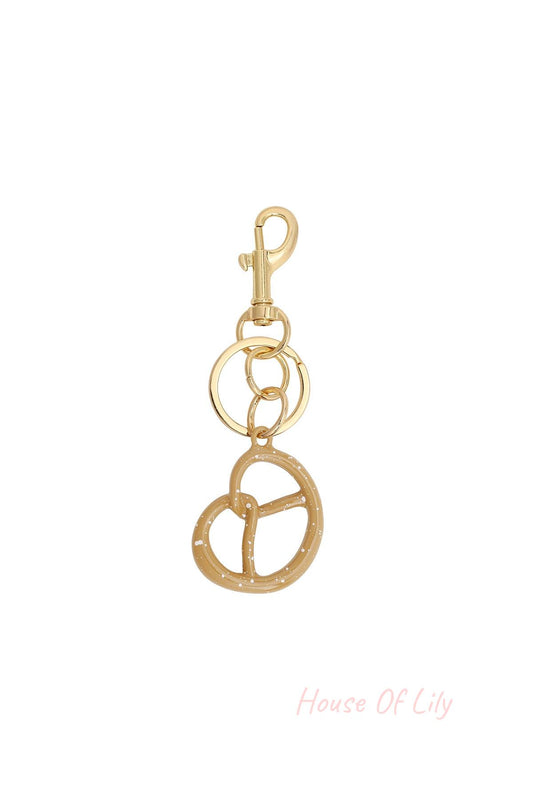 Close-up of a beige pretzel-shaped bag charm with white speckles hanging from gold keyrings and a gold clasp on a white background