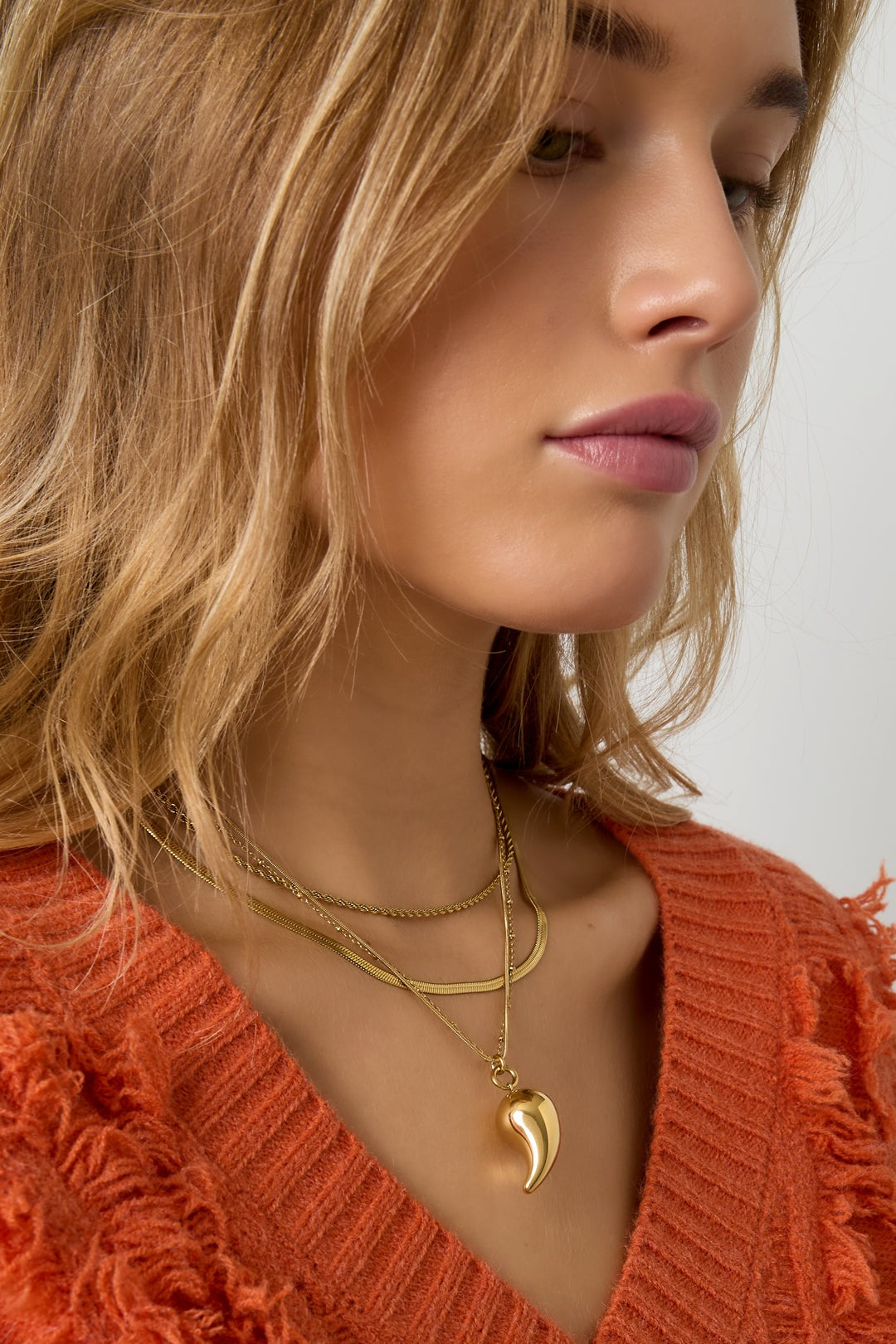 Close-up side view of a woman wearing layered gold necklaces with a droplet pendant paired with an orange textured sweater