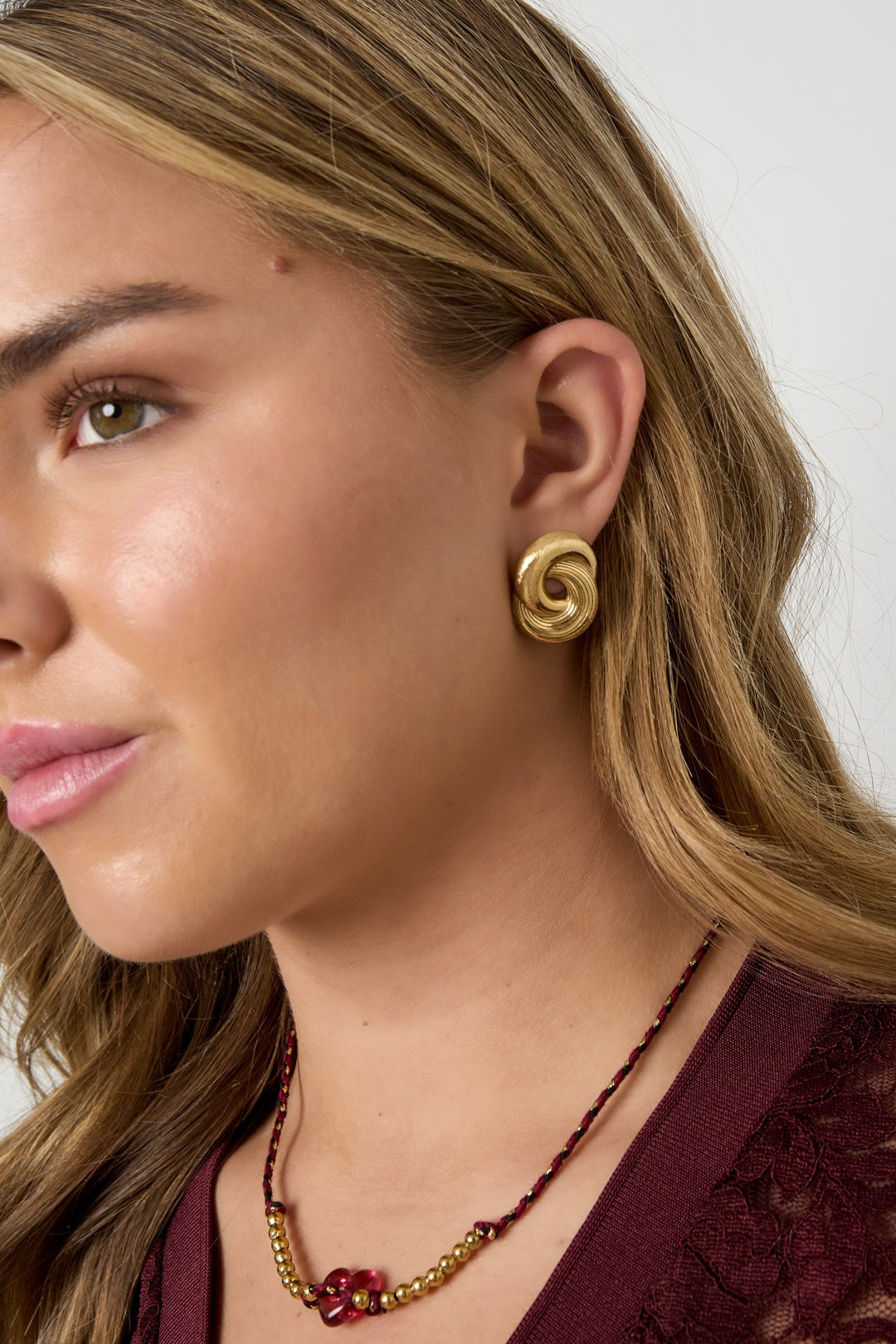 Model wearing gold twist statement stud earrings paired with a burgundy lace top and beaded necklace
