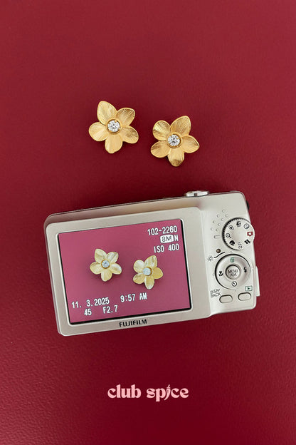 Gold floral zircon stud earrings displayed next to a camera on a red surface with club spice logo