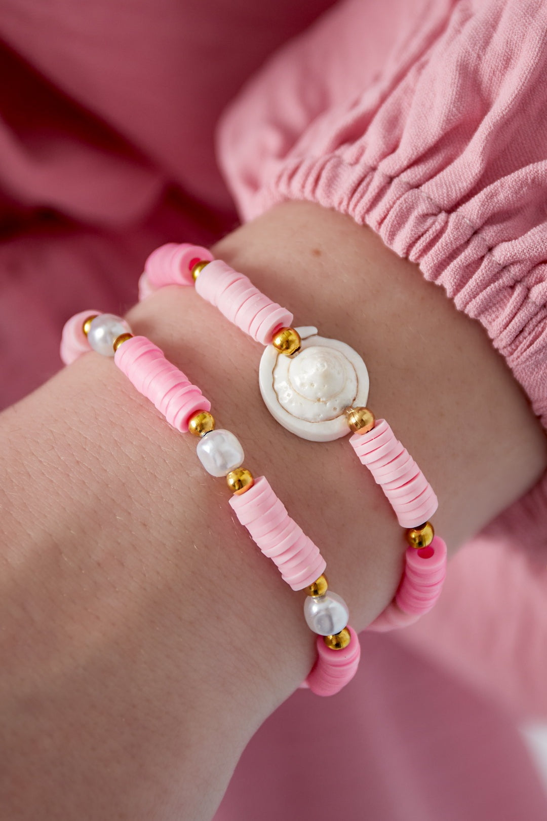 Close-up of two pastel pink shell charm beaded bracelets worn on wrist with pink sleeve
