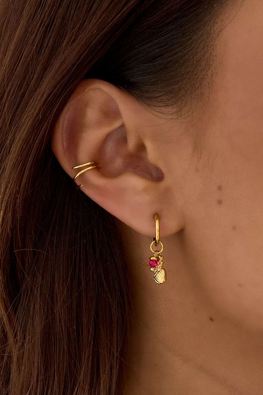 Gold hoop earring with a small gold pineapple charm and a vibrant pink crystal drop, worn on an earlobe.