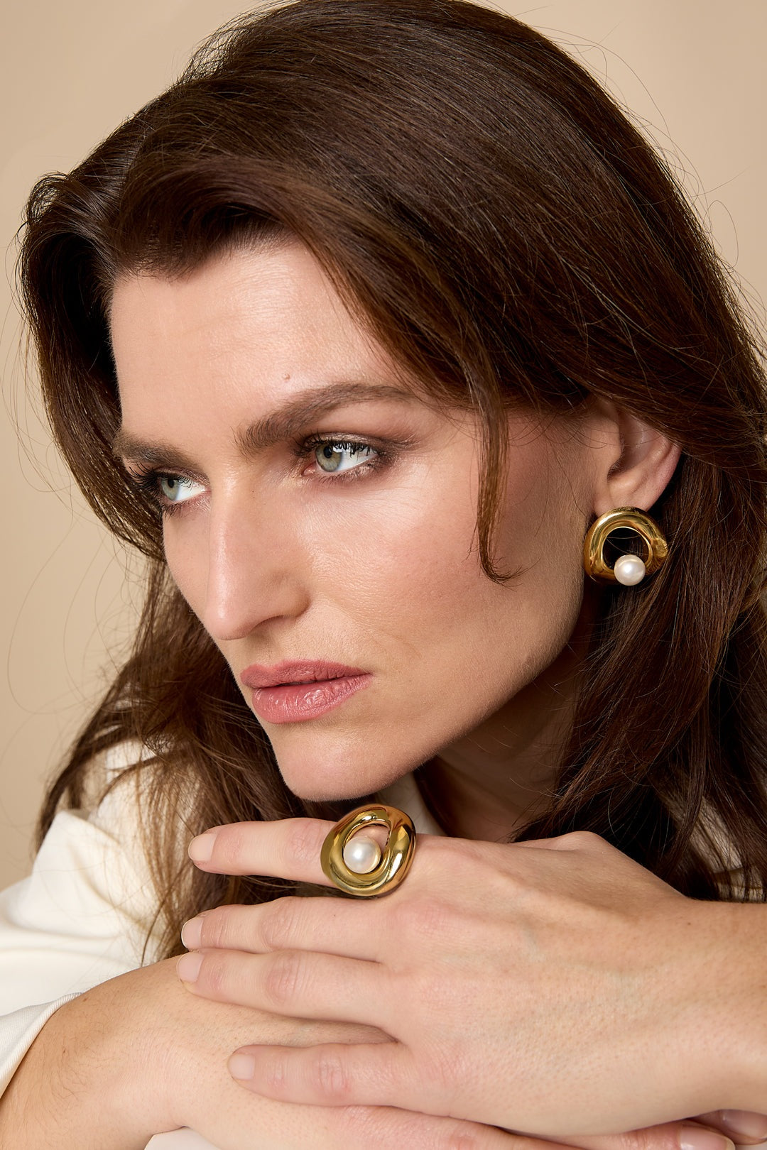 Woman wearing matching gold sculpted pearl earrings and ring, with soft natural makeup and neutral background