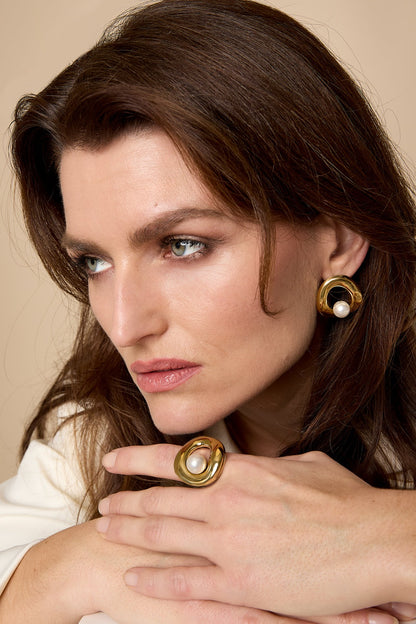 Woman wearing matching gold sculpted pearl earrings and ring, with soft natural makeup and neutral background