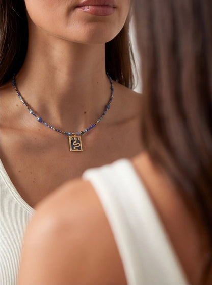 Blue beaded necklace featuring a gold-toned rectangular pendant with a stylized snake design on a blue enamel background.