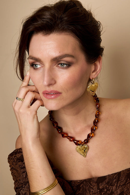 Woman wearing amber beaded necklace with gold heart pendant and matching heart earring, styled with brown off-shoulder top