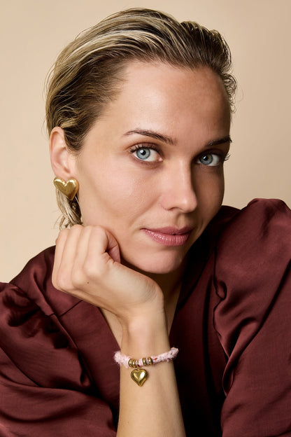 Woman wearing a pink knitted heart charm bracelet and matching gold heart earrings, posing with hand near face