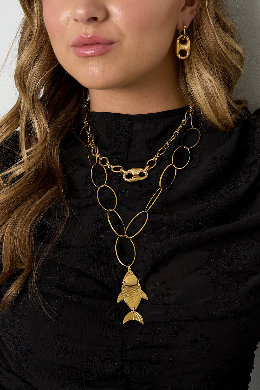 Close-up of woman wearing gold link chain necklace with oval centerpiece and layered gold jewelry over black top