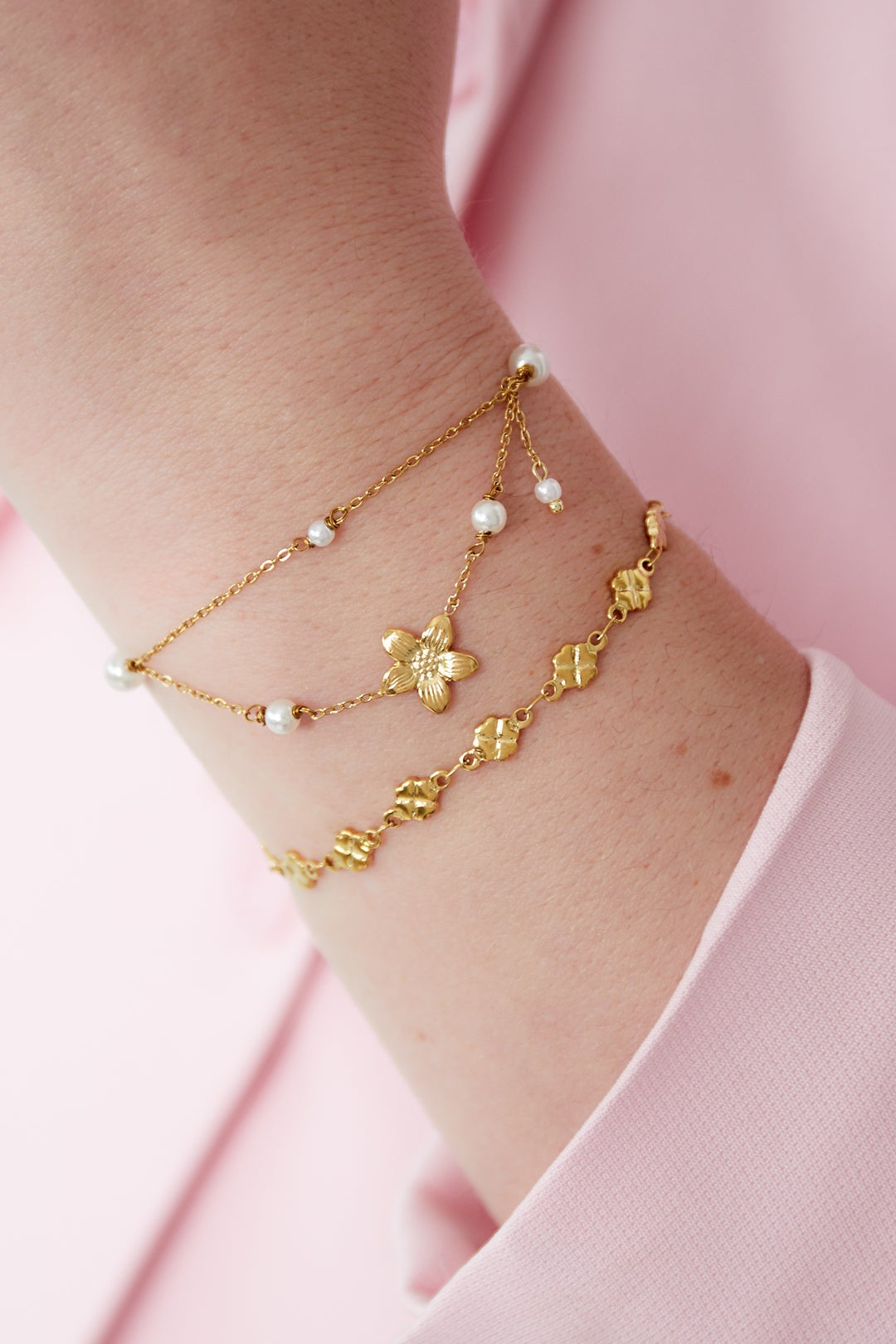 Woman's wrist wearing a layered gold double layer bracelet with flower charm and another floral bracelet, styled with pink sleeve