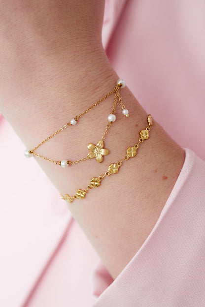 Woman's wrist wearing a layered gold double layer bracelet with flower charm and another floral bracelet, styled with pink sleeve