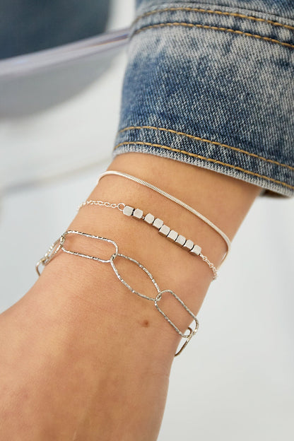 Close-up of silver-tone dainty cube chain bracelet layered with two other bracelets on wrist wearing denim sleeve