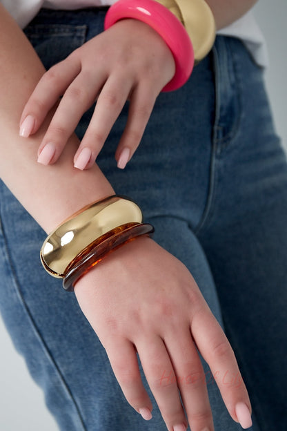 Woman wearing a chunky gold hinged bangle along with bright pink and amber-colored bangles, paired with casual denim jeans