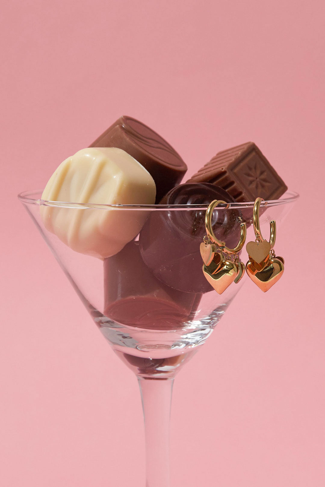 Gold heart charm drop earrings hanging on a clear glass filled with chocolates against pink background
