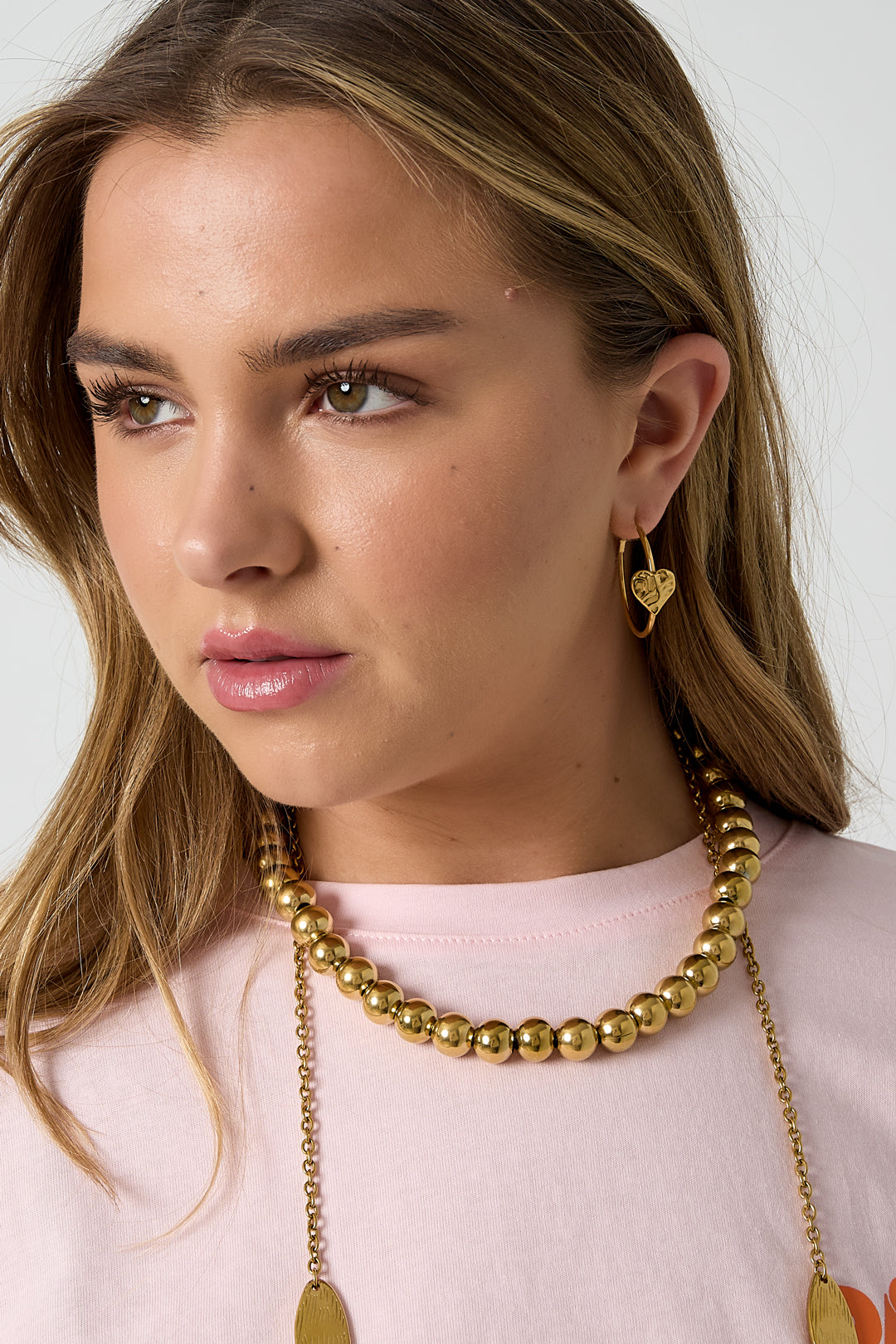 Close-up of woman wearing gold textured heart hoop earrings with gold bead necklace and pink top