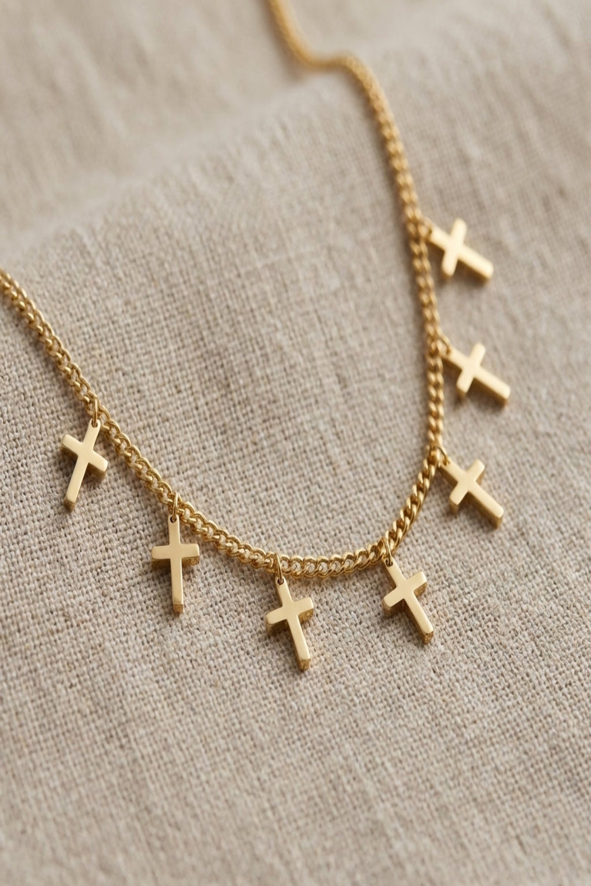 Close-up of delicate seven-cross charm necklace with fine chain, polished silver or gold finish, on neutral textured background.