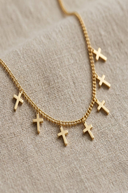 Close-up of delicate seven-cross charm necklace with fine chain, polished silver or gold finish, on neutral textured background.