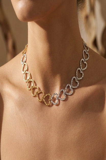 Close-up of a delicate dual-tone gold and silver heart link chain around a model's neck, highlighting hammered texture and natural lighting.