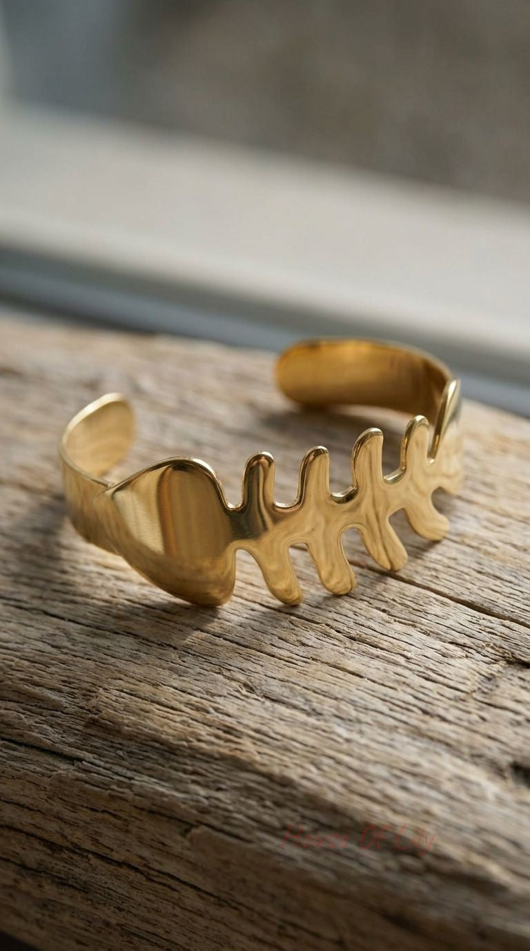 Close-up of a small gold fishbone cuff bangle with detailed cut-out design on textured wooden surface.