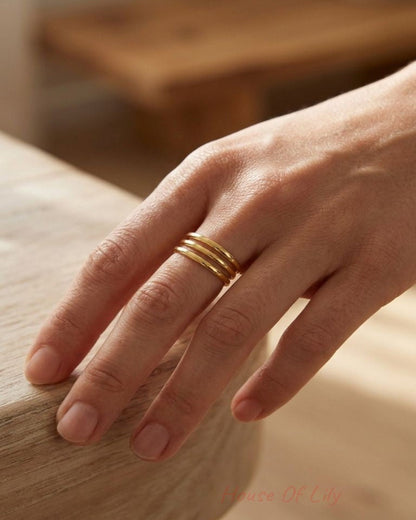 Close-up of the 'Triple Layer Gold' ring on a natural skin tone hand, showing three parallel stainless steel bands with a gold-tone finish on a textured wooden surface.