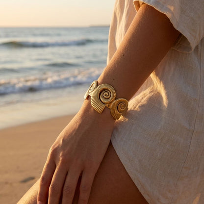 Model wearing Coastal Spiral Cuff on wrist against beach backdrop with golden hour lighting and seaside scenery.