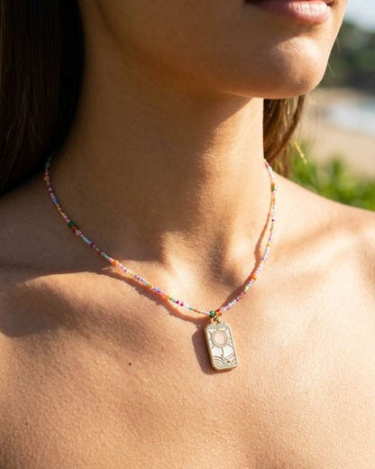 Close-up of colourful sun charm necklace with vibrant beads and golden pendant on model's collarbone outdoors.