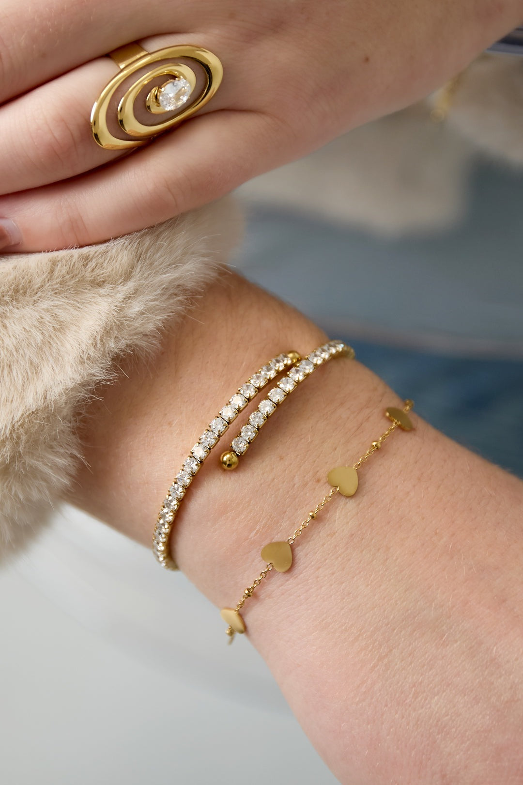 A woman’s wrist adorned with the gold-tone sparkling crystal cuff bracelet, a gold ring with spiral design and gemstone, and a delicate gold heart chain bracelet