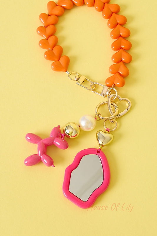 Bright pink mirror charm featuring gold heart and pearl accents attached to an orange heart-linked strap on yellow background